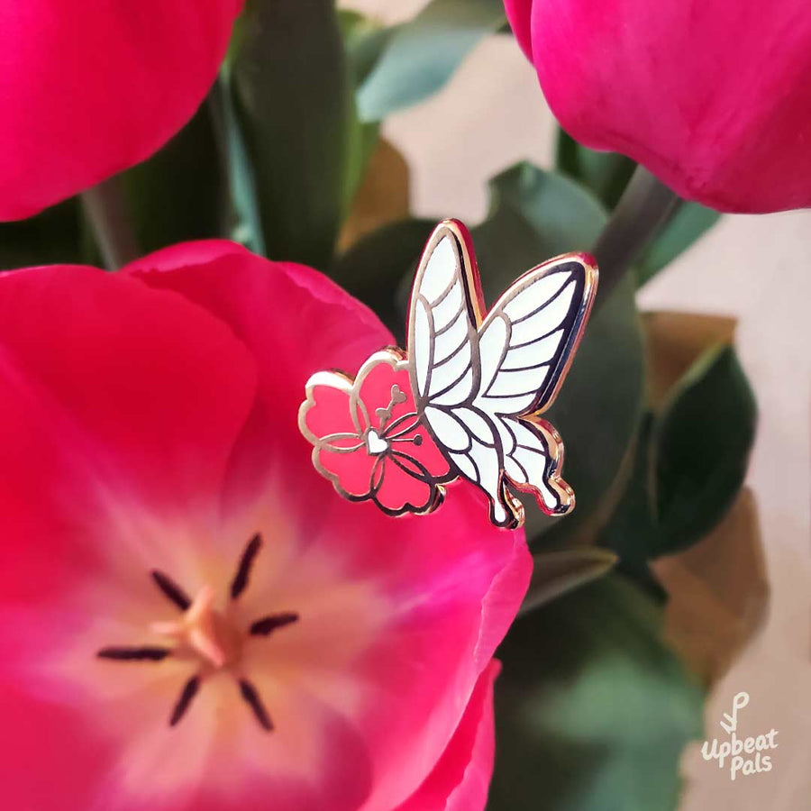 A gold plated enamel pin featuring a white butterfly landing on a red flower. This lapel pin is shown on top of a potted tulip plant.