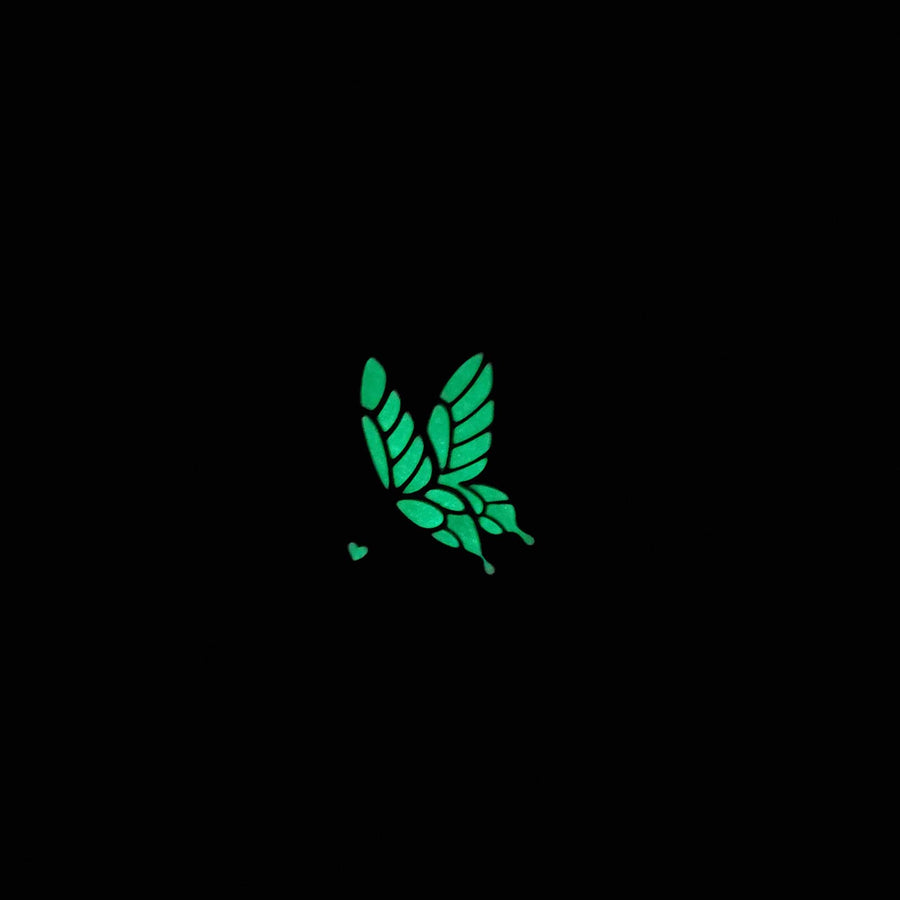 A gold plated enamel pin featuring a white butterfly landing on a red flower. This lapel pin in complete darkness to highlight that it glows in the dark with a neon green color.