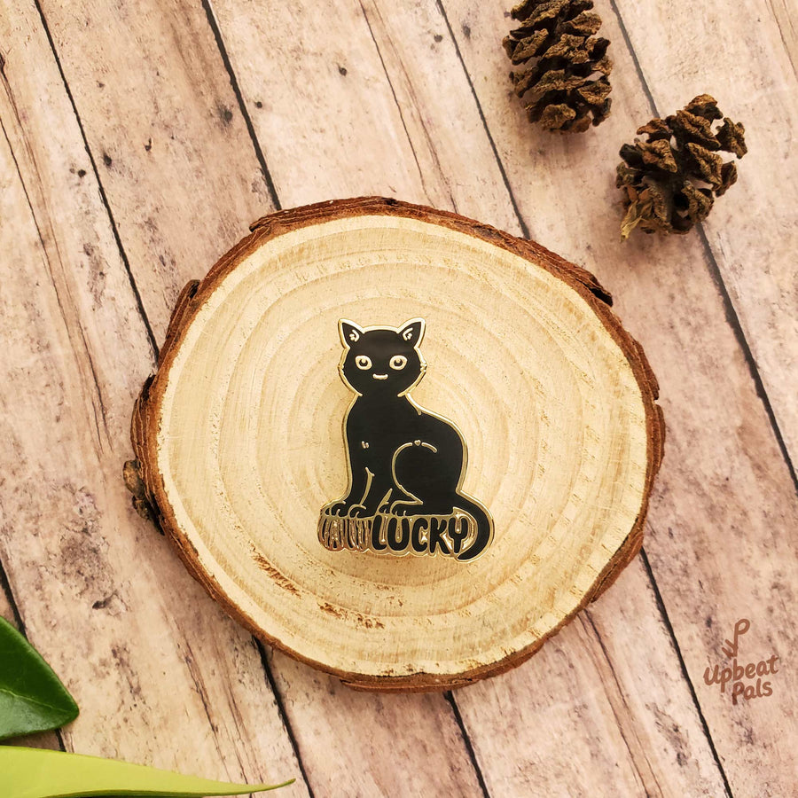 A gold plated enamel pin of a cat perched on top of the word lucky, in black letters. The cat's forepaws rest on the letters U and N, to transform the word from unlucky into lucky. This lapel pin sits on top of a decorative wood stump.