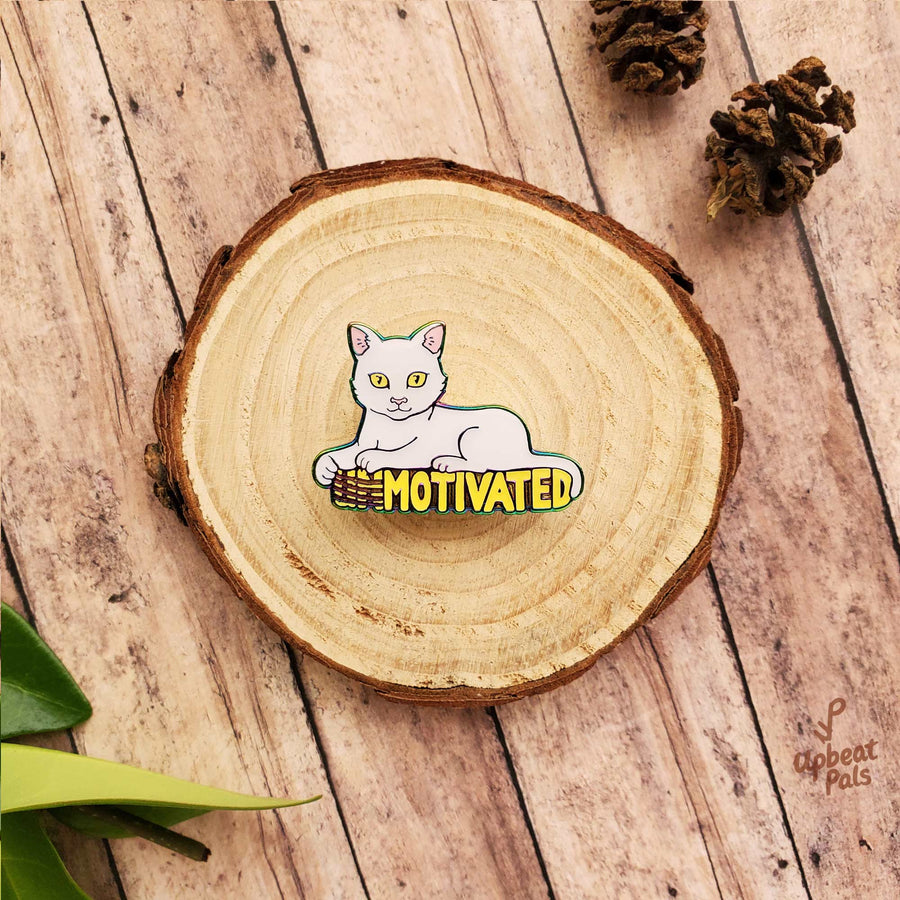 A rainbow metal enamel pin of a white cat sitting on top of the word motivated, in yellow letters. The cat's forepaws have scratched off the U and N, to transform the word from unmotivated into motivated. This lapel pin sits on top of a decorative wood stump.