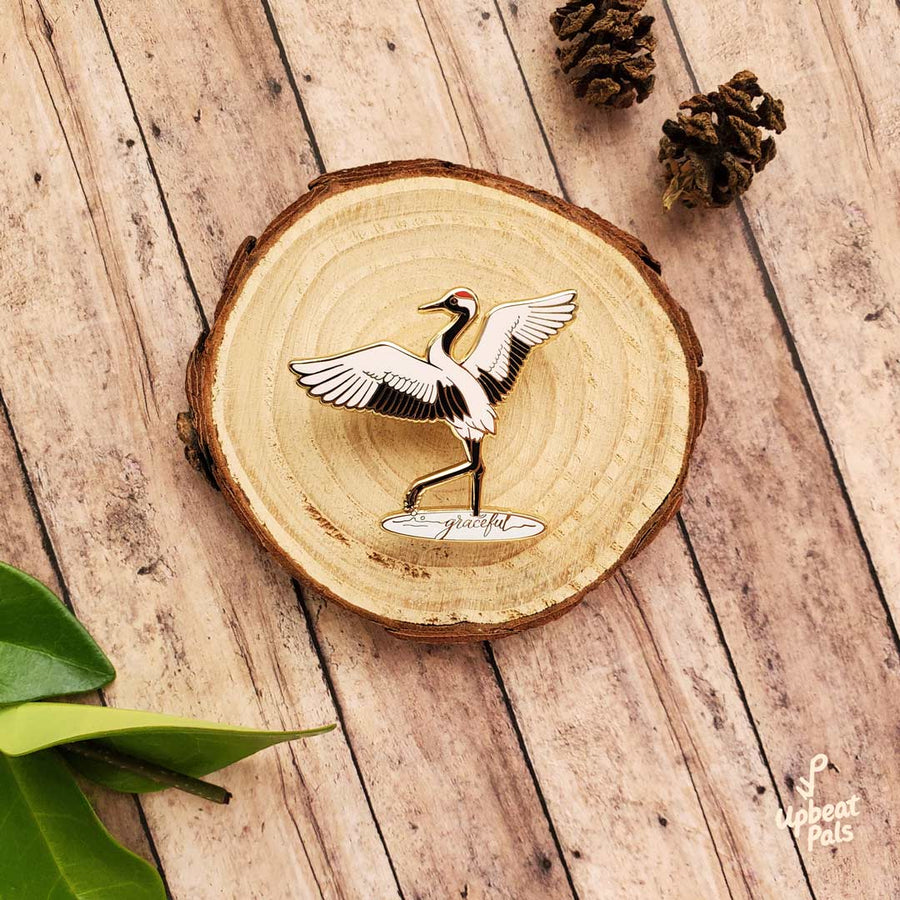 A gold plated enamel pin of crane with wings outstretched, lifting one leg out of a pool of water. In the pool of water is the word graceful, written in cursive. This lapel pin is displayed on a decorative wood stump.