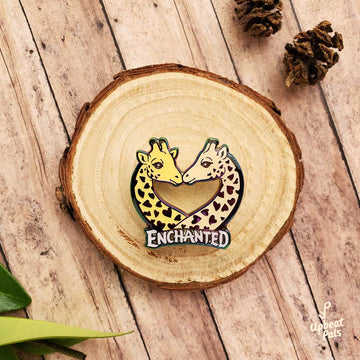 A rainbow metal enamel pin of two giraffes busts, with their faces nuzzling. The word enchanted appears at the bottom of the busts. This lapel pin sits on a decorative tree stump.