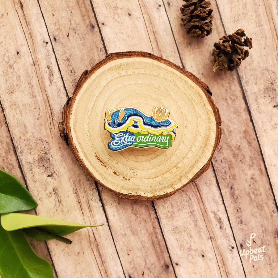 A glitter enamel pin of a seaslug nudibranch on top of the word extraordinary. This lapel pin of chromodoris annae sits on top of a decorative tree stump.