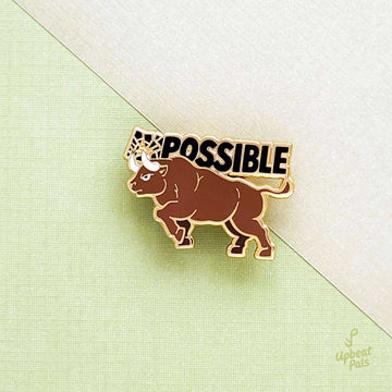 A gold plated enamel pin of a bull goring the I and M in the word impossible, transforming it into possible. The word possible appears in black letters above the bull, who is charging slightly upward. This lapel pin sits two pieces of intersecting paper.