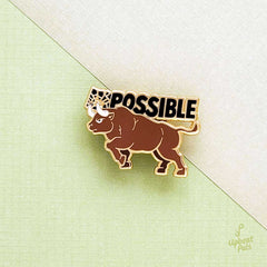 A gold plated enamel pin of a bull goring the I and M in the word impossible, transforming it into possible. The word possible appears in black letters above the bull, who is charging slightly upward. This lapel pin sits two pieces of intersecting paper.