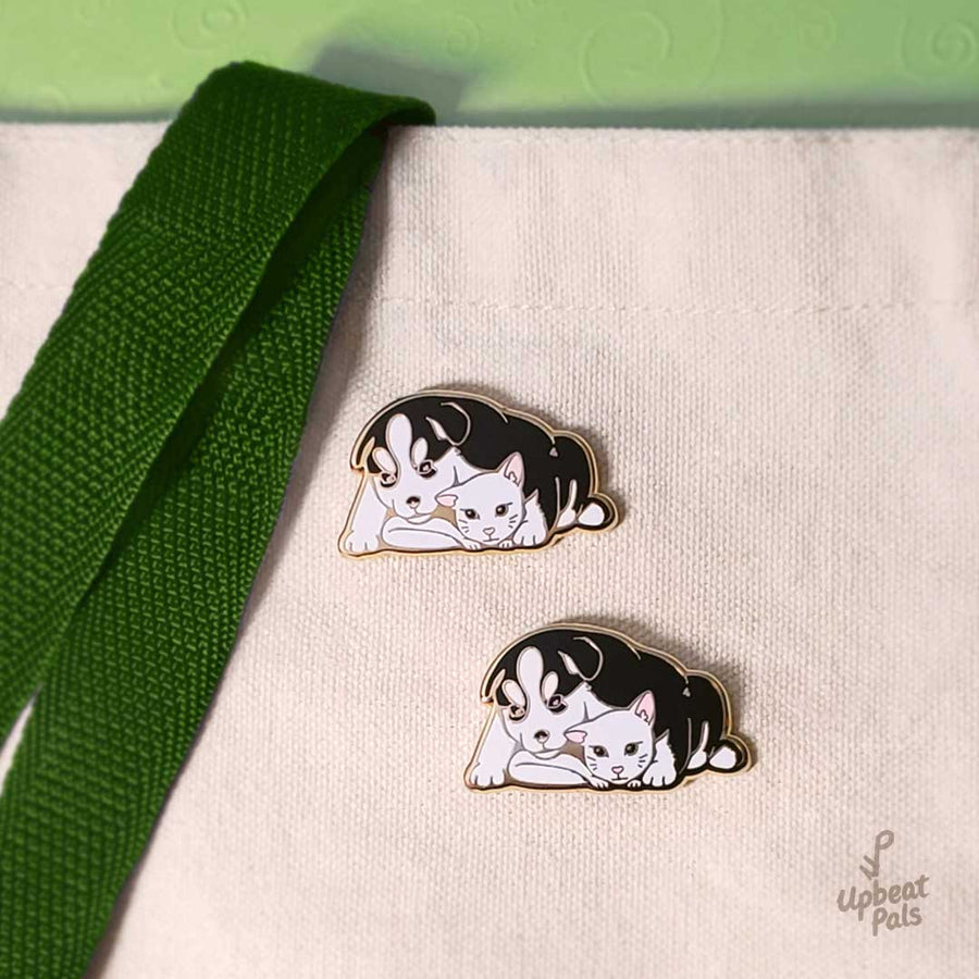 Two gold plated enamel pins of a husky dog and white cat cuddling and looking at each other. These lapel pins are attached to a reusable tote bag.