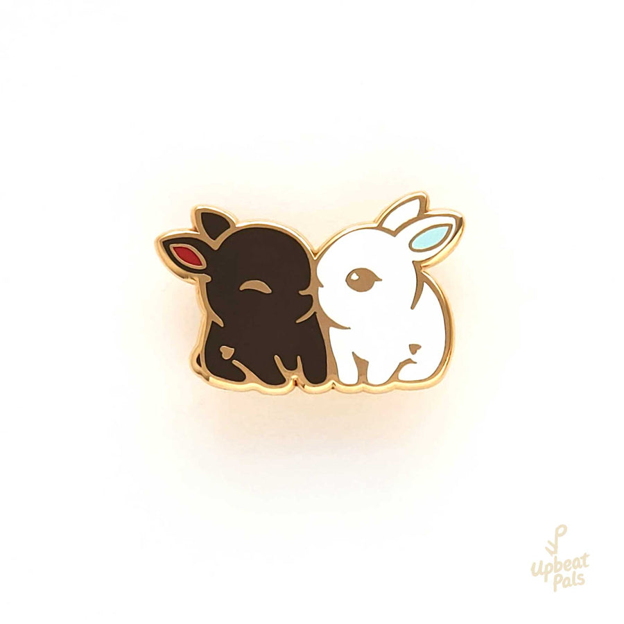 An enamel pin of two cute bunny rabbits snuggling. One is chocolate brown with red ears and the other is pure white with light blue ears. This lapel pin is displayed on a white background.