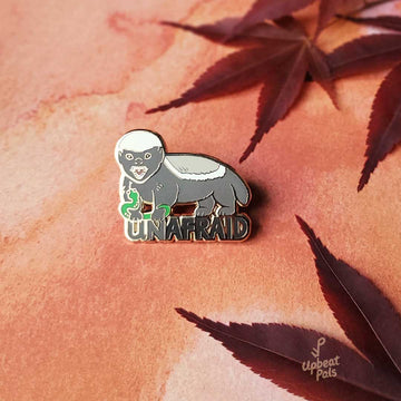 An enamel pin of brave honey badger wrestling a green snake, on top the word Unafraid. This lapel pin features gold plating technology and is shown on a red decorative background with leaves.