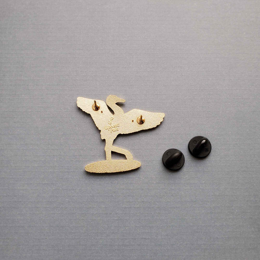 The back of a gold plated enamel pin of crane with wings outstretched, lifting one leg out of a pool of water. In the pool of water is the word graceful, written in cursive. This lapel pin sits on a textured gray background.