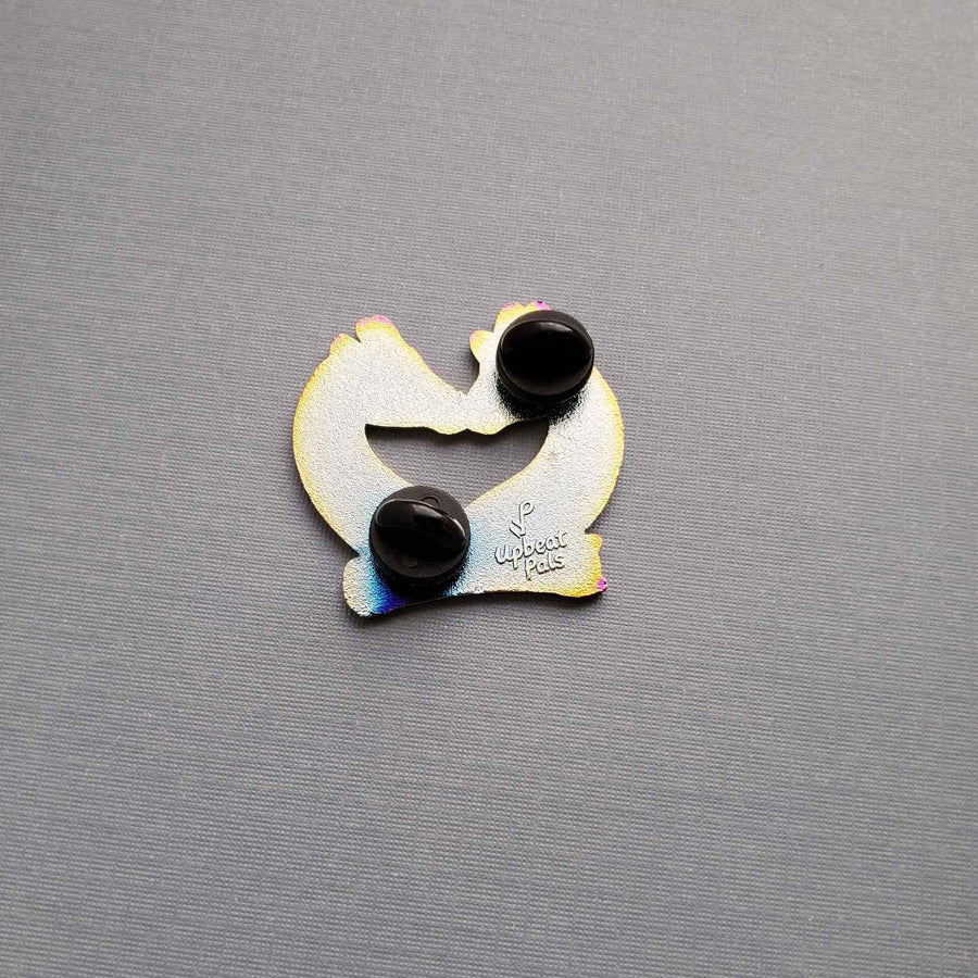 The back of a rainbow metal enamel pin of two giraffes busts, with their faces nuzzling. The word enchanted appears at the bottom of the busts. This pin sits on top of a gray textured background.