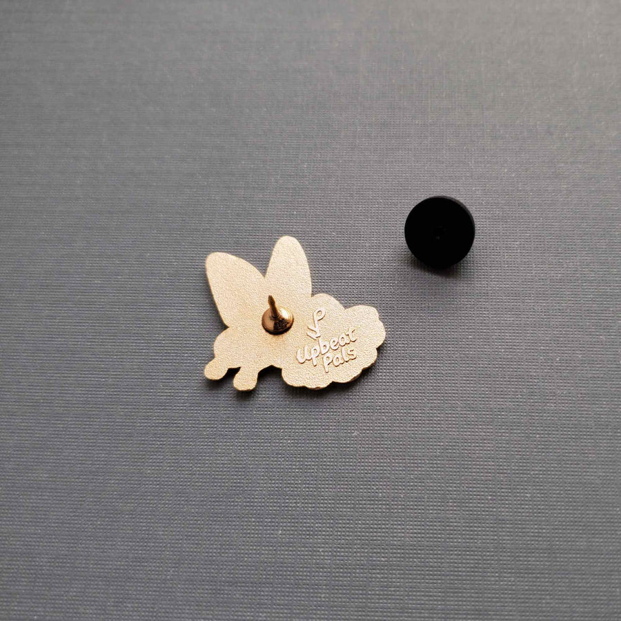 The back of a gold plated enamel pin featuring a white butterfly landing on a red flower. This lapel pin is shown with its pin backing off, revealing the pin post.