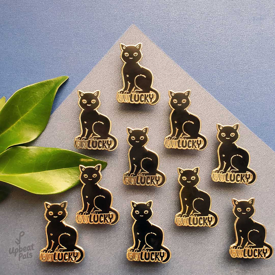 Ten gold plated enamel pins of a cat perched on top of the word lucky, in black letters. The cat's forepaws rest on the letters U and N, to transform the word from unlucky into lucky. These lapel pins are shown on top of decorative colored paper.