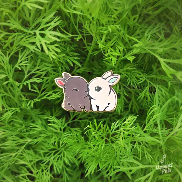 An enamel pin of two cute bunny rabbits snuggling. One is chocolate brown with red ears and the other is pure white with light blue ears. This lapel pin is displayed with a bed of dill plants in the background.