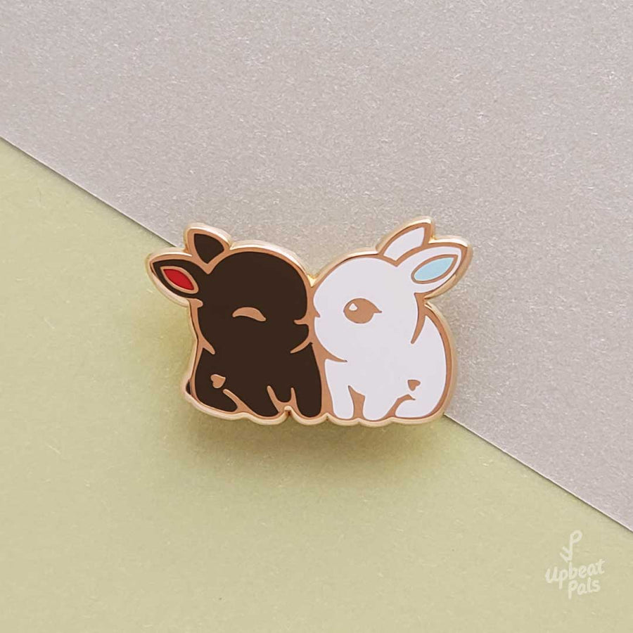 An enamel pin of two cute bunny rabbits snuggling. One is chocolate brown with red ears and the other is pure white with light blue ears. This lapel pin is displayed on a two color background.