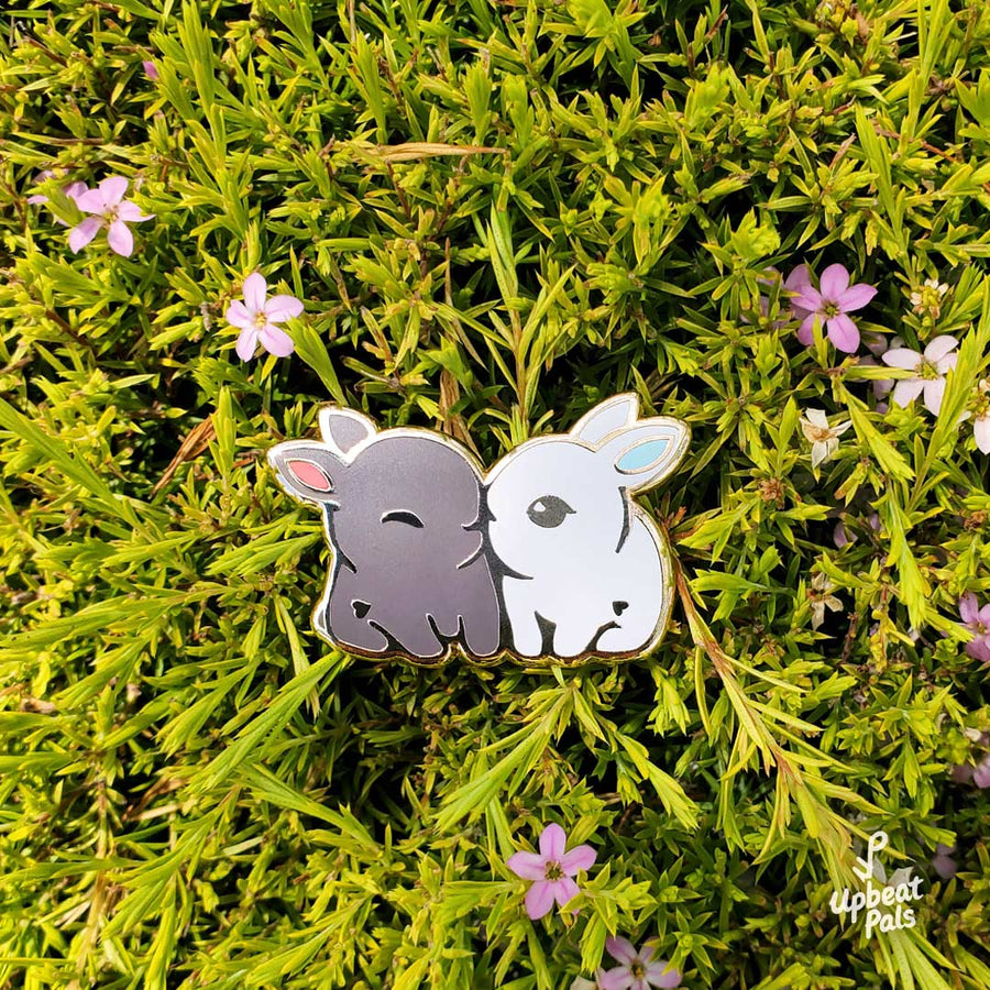 An enamel pin of two cute bunny rabbits snuggling. One is chocolate brown with red ears and the other is pure white with light blue ears. This lapel pin is displayed on top of a plant with light purple flowers.