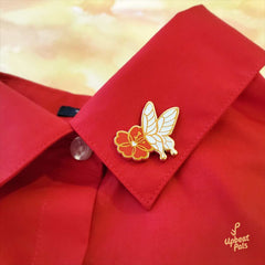 A gold plated enamel pin featuring a white butterfly landing on a red flower. This lapel pin is shown affixed on the neck collar of a red dress shirt.