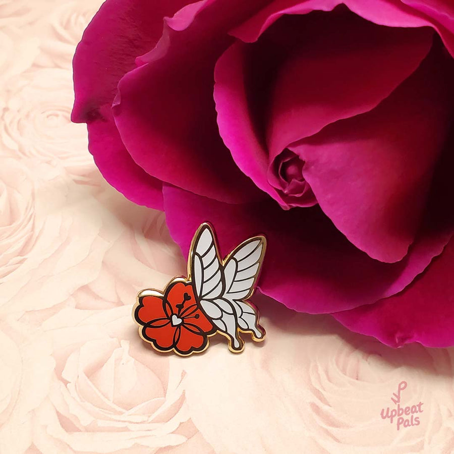 A gold plated enamel pin featuring a white butterfly landing on a red flower. This lapel pin is next to a red rose with a decorative floral background.