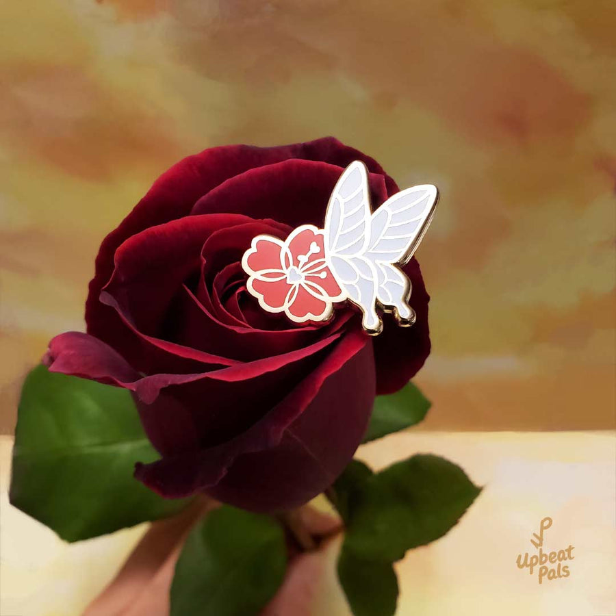 A gold plated enamel pin featuring a white butterfly landing on a red flower. This lapel pin is shown on top of a crimson red rose.