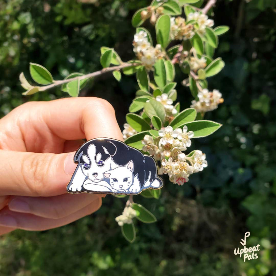 A gold plated enamel pin of a husky dog and white cat cuddling and looking at each other. This lapel pin is beind held in front of a branch of white cluster flowers.