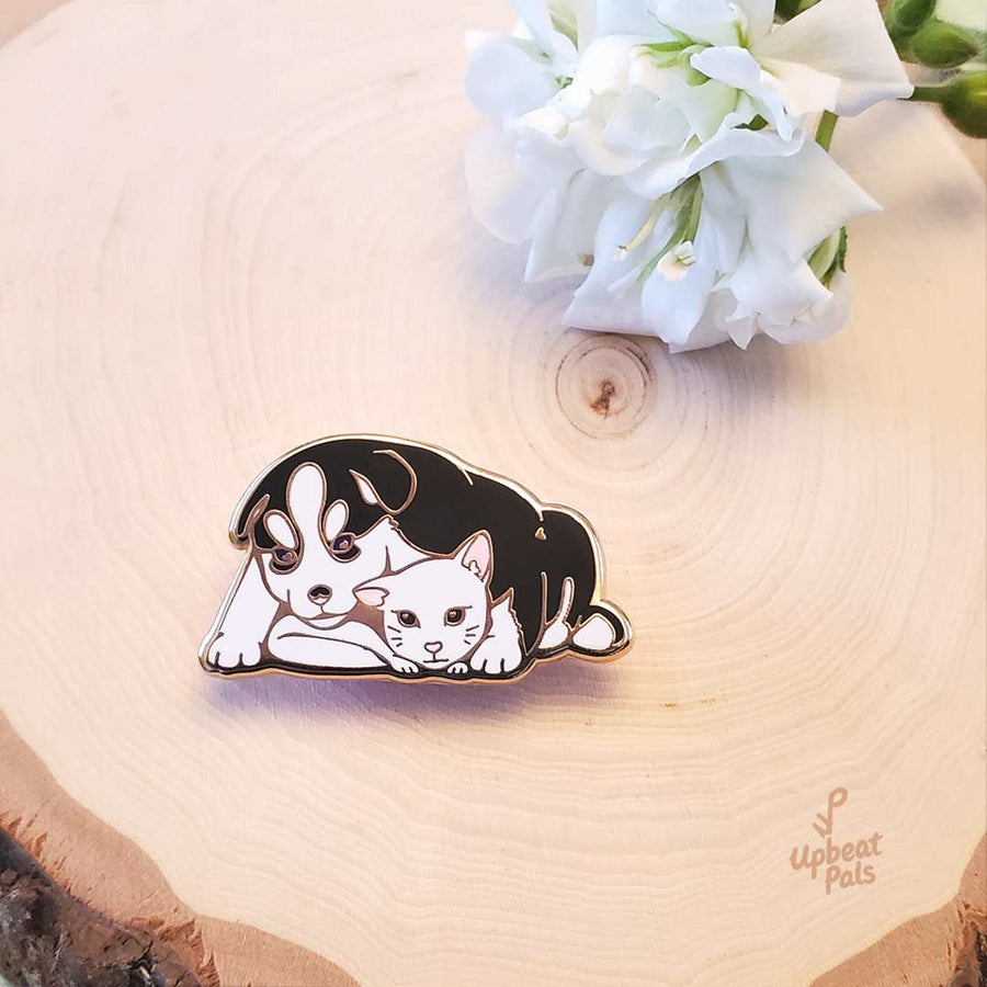 A gold plated enamel pin of a husky dog and white cat cuddling and looking at each other. This lapel pin is resting on a decorative wooden stump with white flowers placed near the pin.