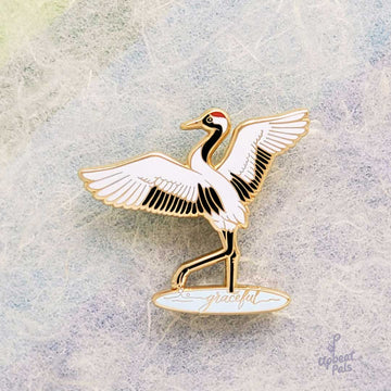 A gold plated enamel pin of crane with wings outstretched, lifting one leg out of a pool of water. In the pool of water is the word graceful, written in cursive. This lapel pin is displayed in front of textured multicolor paper.