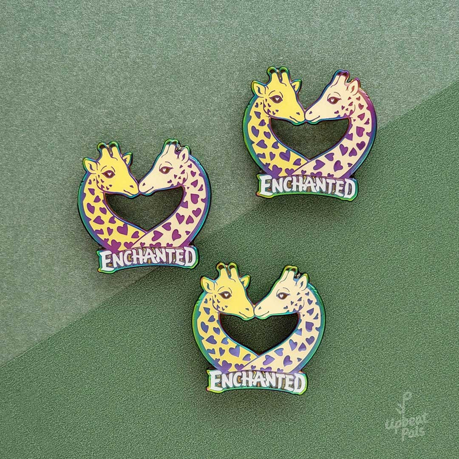 Three rainbow metal enamel pins of two giraffes busts, with their faces nuzzling. The word enchanted appears at the bottom of the busts. These lapel pins sit on top of two-tone green decorative paper.