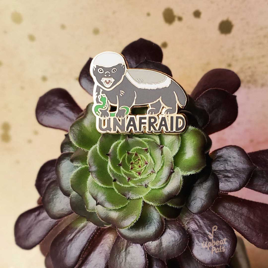 A enamel pin of brave honey badger wrestling a green snake, on top the word Unafraid. This lapel pin was placed on a plant. 