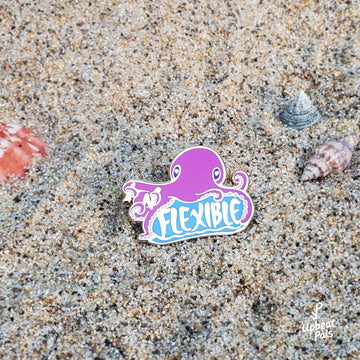 A gold plated enamel pin of a purple octopus sitting on top of the word flexible. In its tentacles are the letters I and N, which transform the word from inflexible to flexible. This lapel pin rests on a beach sand background.