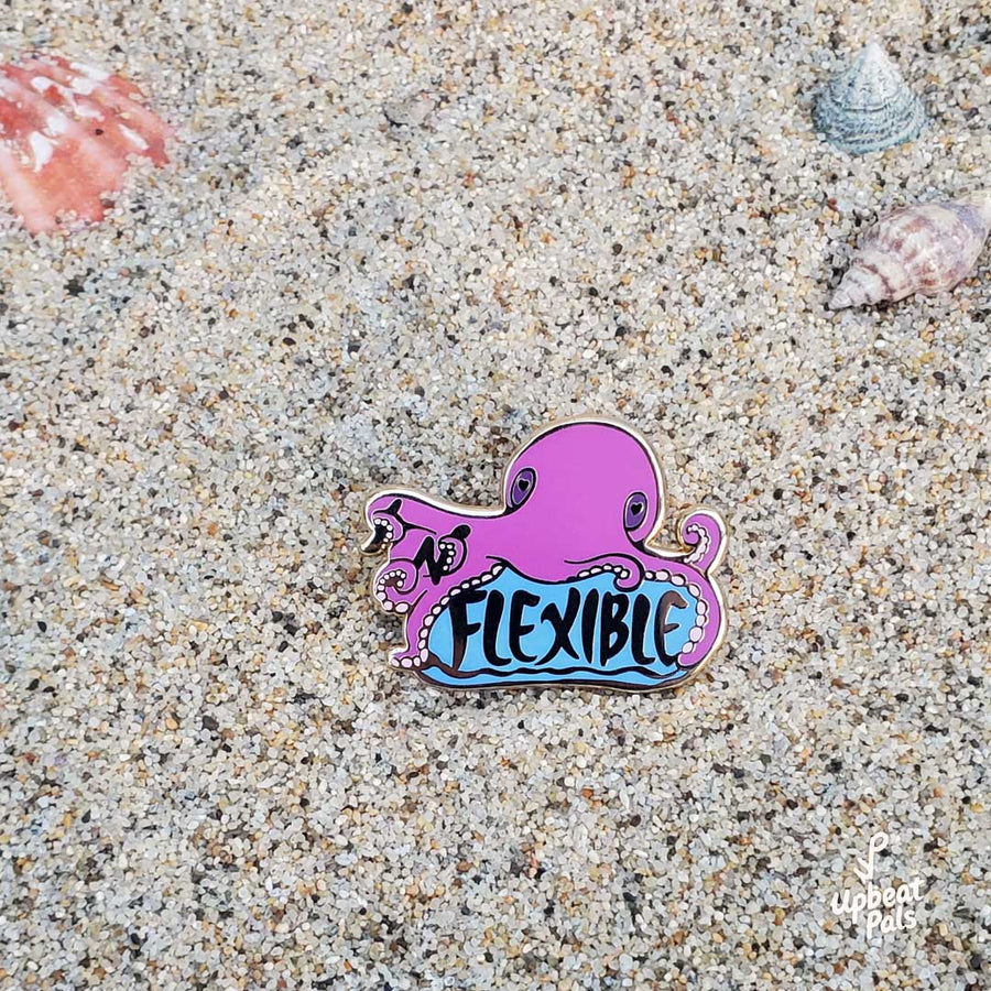 A gold plated enamel pin of a purple octopus sitting on top of the word flexible. In its tentacles are the letters I and N, which transform the word from inflexible to flexible. This lapel pin rests on a beach sand background.