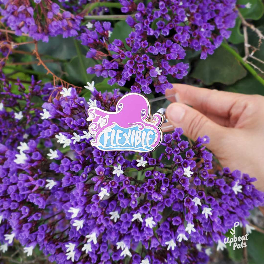 A gold plated enamel pin of a purple octopus sitting on top of the word flexible. In its tentacles are the letters I and N, which transform the word from inflexible to flexible. This lapel pin is being held in front of a bunch of minature purple flowers.