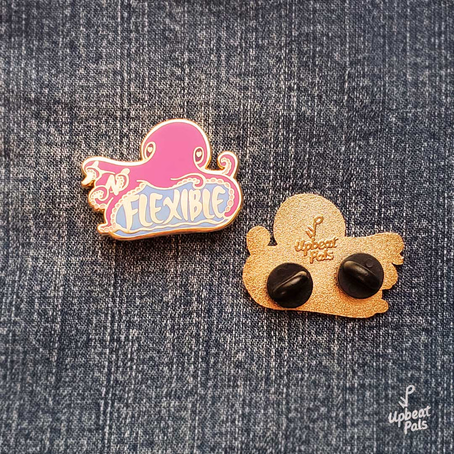 Two gold plated enamel pins of a purple octopus sitting on top of the word flexible. In its tentacles are the letters I and N, which transform the word from inflexible to flexible. One lapel pin faces front while the other faces back, and both sit on textured background.