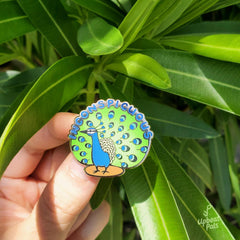 A gold plated enamel pin of a peacock standing on a patch of brown dirt. The word inconspicuous appears around the edge of the tailfeathers, and the "in" in inconspicuous appears pecked out by the peacock. This lapel pin is beind held in front of a green plant.