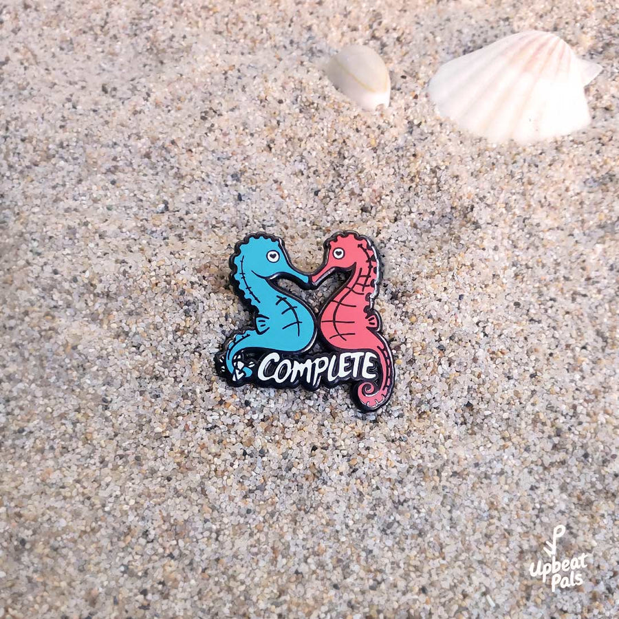 A black nickel plated pin of two seahorses, a blue and red one, joined at their snouts. The word complete runs across the pin, below their bellies in white letters, and the blue seahorse has wrapped up the I and N in its tail to transform this word from incomplete to complete. This lapel pin a beach sand background.