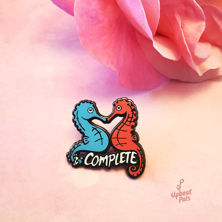 A black nickel plated pin of two seahorses, a blue and red one, joined at their snouts. The word complete runs across the pin, below their bellies in white letters, and the blue seahorse has wrapped up the I and N in its tail to transform this word from incomplete to complete. This lapel pin sits on a floral background.