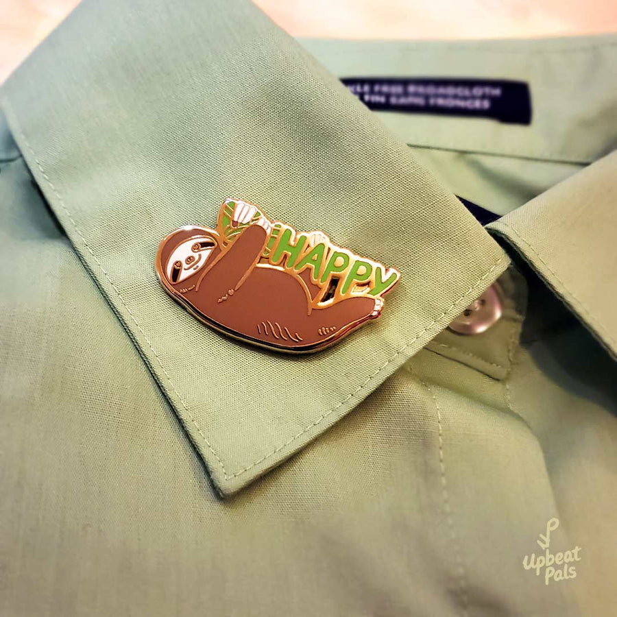 A gold plated enamel pin of a smiling sloth hanging onto the word happy in green letters. The sloth is covering up the letters U and N, to transform the word from unhappy into happy. This lapel pin sits on the collar of a green dress shirt.