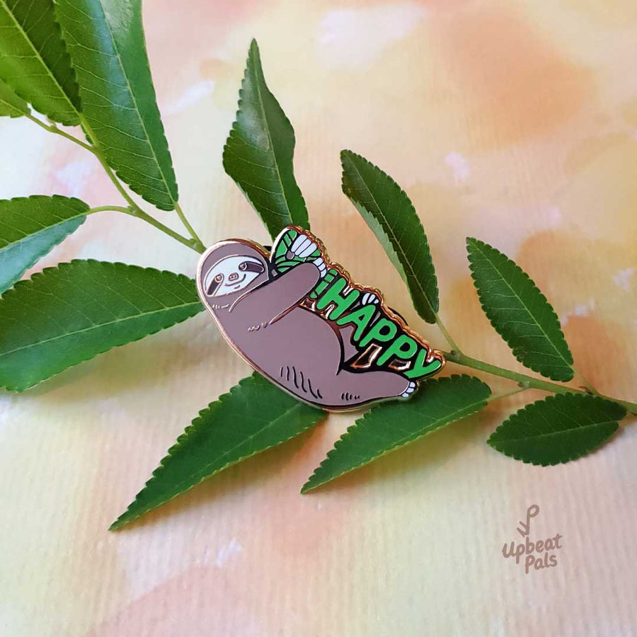 A gold plated enamel pin of a smiling sloth hanging onto the word happy in green letters. The sloth is covering up the letters U and N, to transform the word from unhappy into happy. This lapel pin sits on a plant branch.