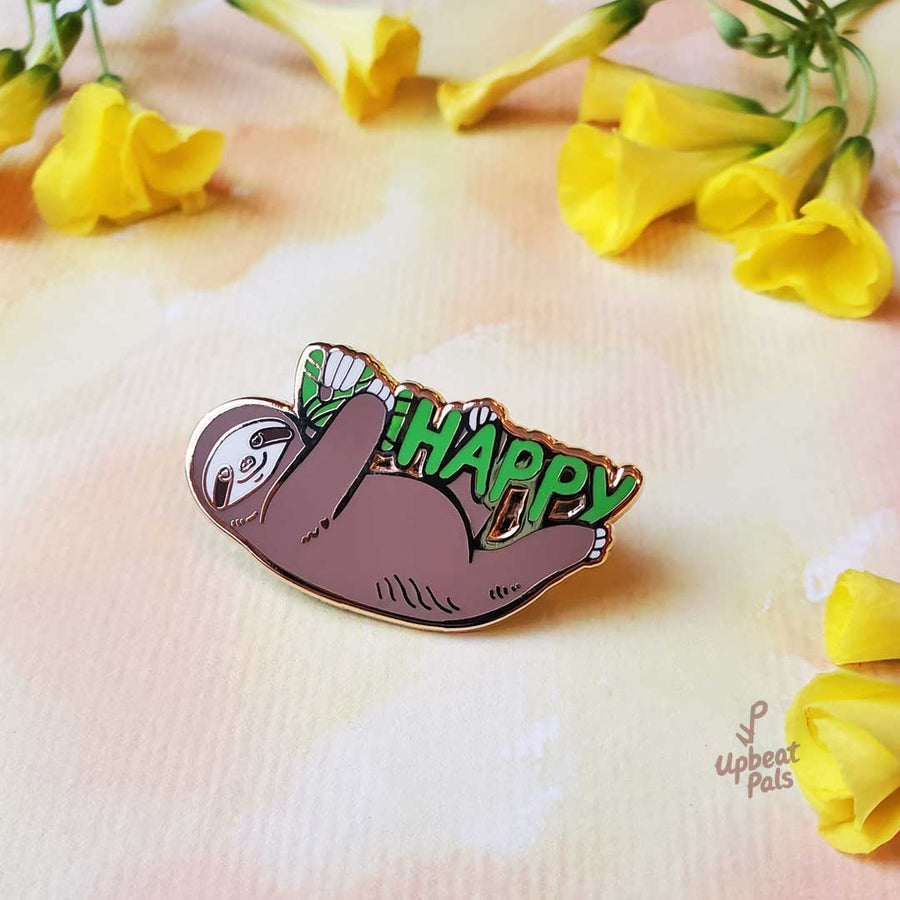 A gold plated enamel pin of a smiling sloth hanging onto the word happy in green letters. The sloth is covering up the letters U and N, to transform the word from unhappy into happy. This lapel pin sits on textured paper surrounded by yellow flowers.