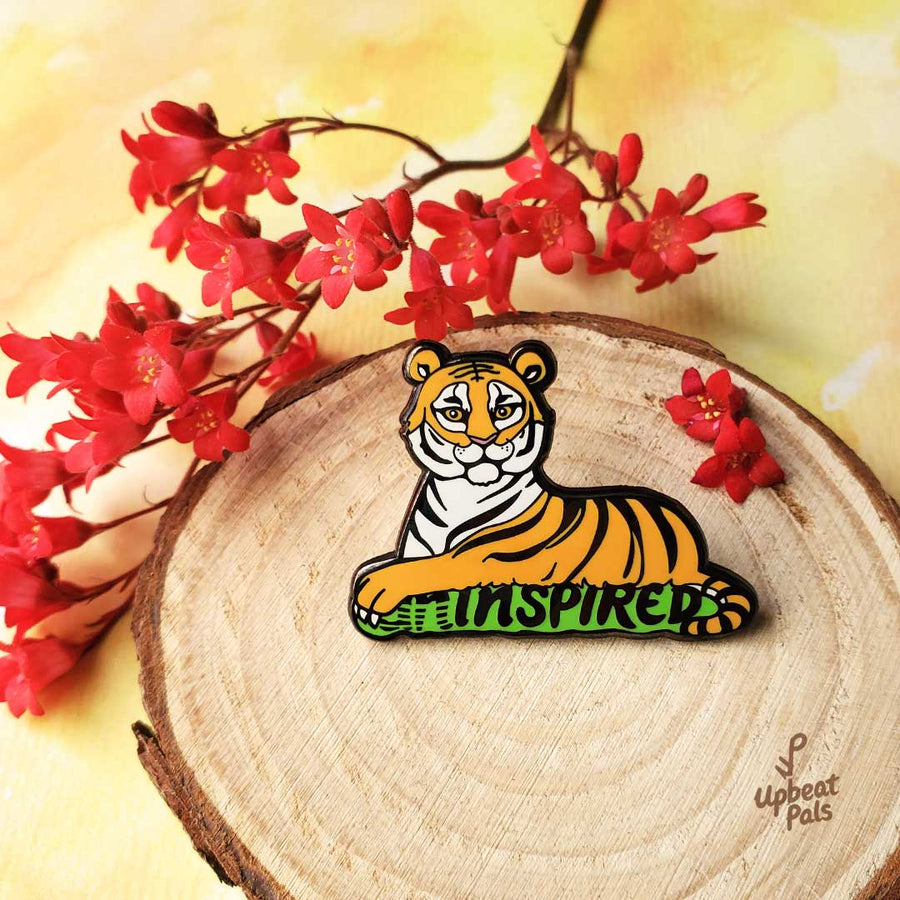 A black nickel plated enamel pin of sitting tiger, resting on top of the word inspired in black letters and a green outline. The tiger's left forepaw is covering the letters U and N, to transform the word from uninspired into inspired. This lapel pin sits on a decorative tree stump.