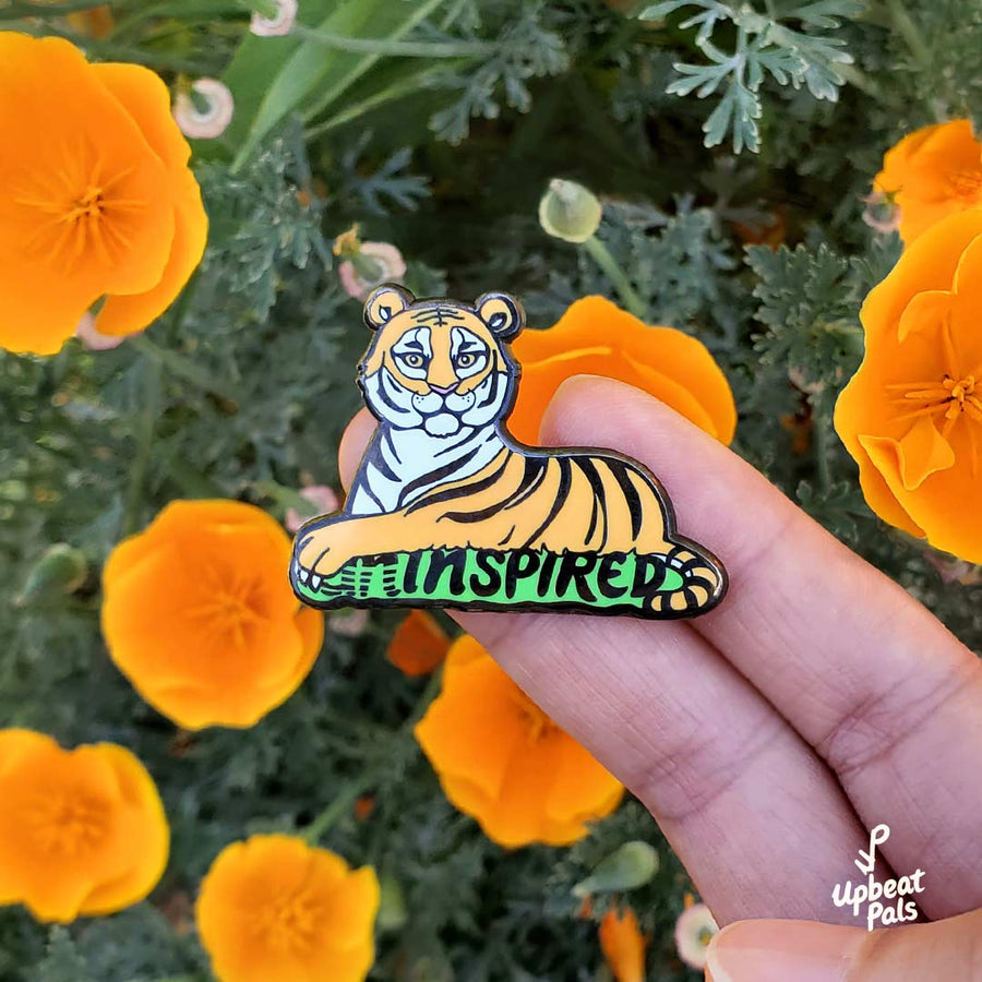 A black nickel plated enamel pin of sitting tiger, resting on top of the word inspired in black letters and a green outline. The tiger's left forepaw is covering the letters U and N, to transform the word from uninspired into inspired. This lapel pin is held in front of orange poppies.