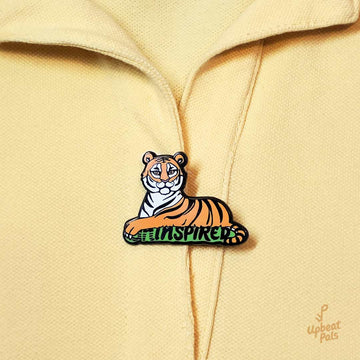 A black nickel plated enamel pin of sitting tiger, resting on top of the word inspired in black letters and a green outline. The tiger's left forepaw is covering the letters U and N, to transform the word from uninspired into inspired. This lapel pin is on top of a yellow polo shirt.