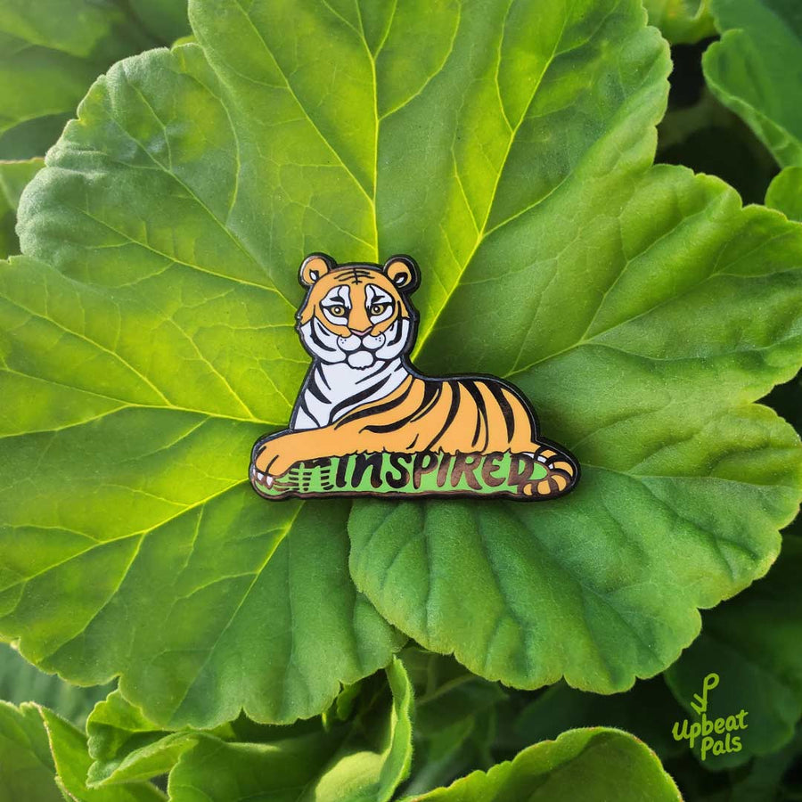 A black nickel plated enamel pin of sitting tiger, resting on top of the word inspired in black letters and a green outline. The tiger's left forepaw is covering the letters U and N, to transform the word from uninspired into inspired. This lapel pin is on top of a large leaf.