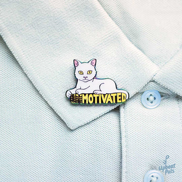 A rainbow metal enamel pin of a white cat sitting on top of the word motivated, in yellow letters. The cat's forepaws have scratched off the U and N, to transform the word from unmotivated into motivated. This lapel pin sits on  the collar of a blue polo shirt.